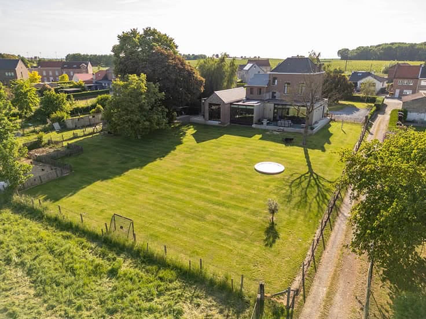 Luchtfoto van het volledige perceel: de gebouwen bovenaan, daaronder een lang aflopend gazon met bomen aan weerszijden en velden erachter.
