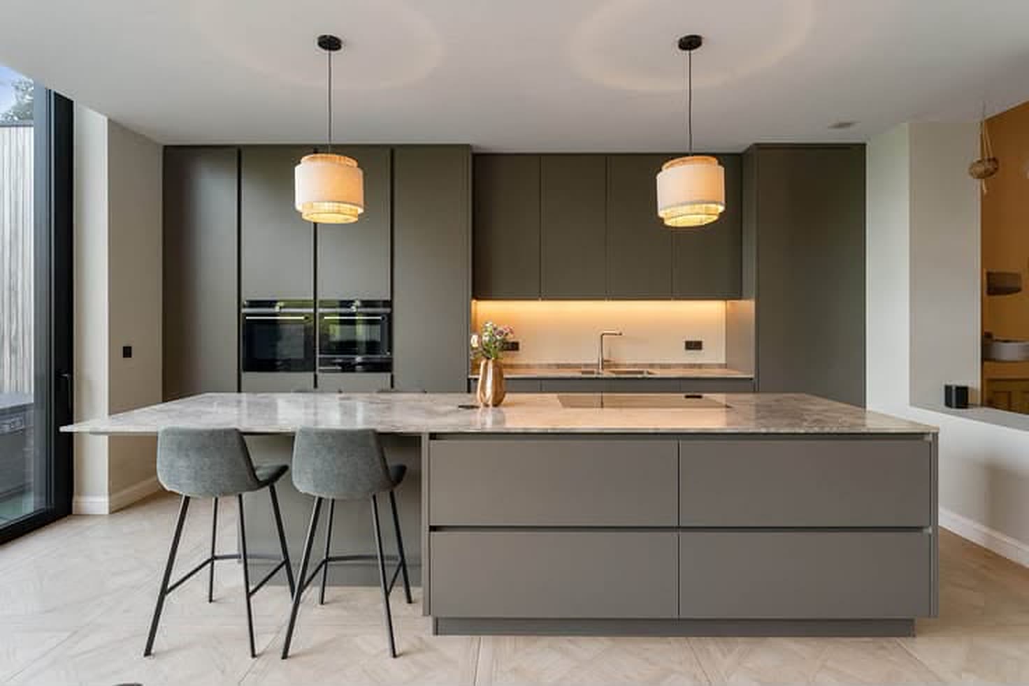 Keuken met groot eiland, twee barkrukken, donkergrijze kasten en ingebouwde ovens.