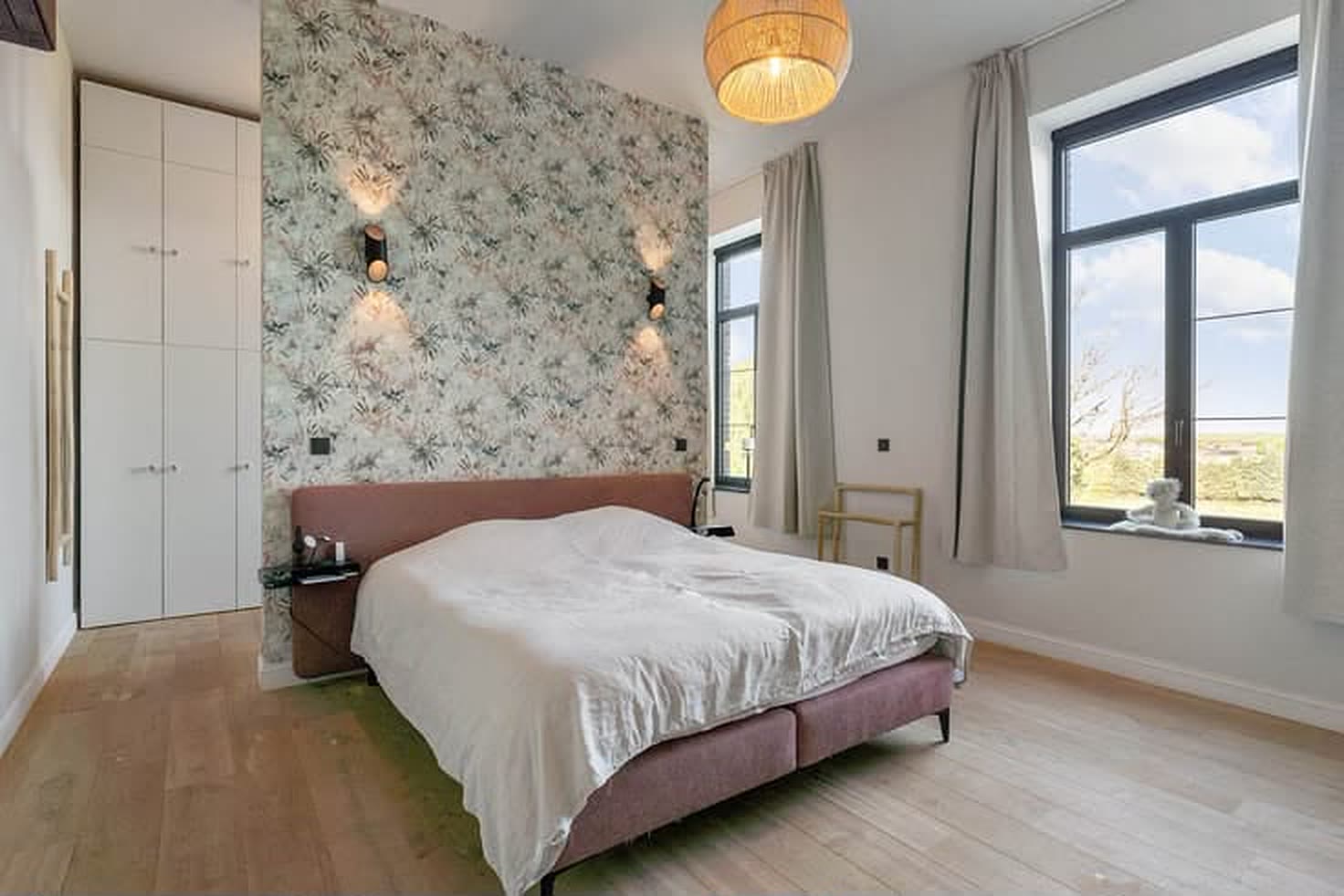 Hoofdslaapkamer met bloemenbehang achter het bed, ingebouwde kasten en een raam op de velden.
