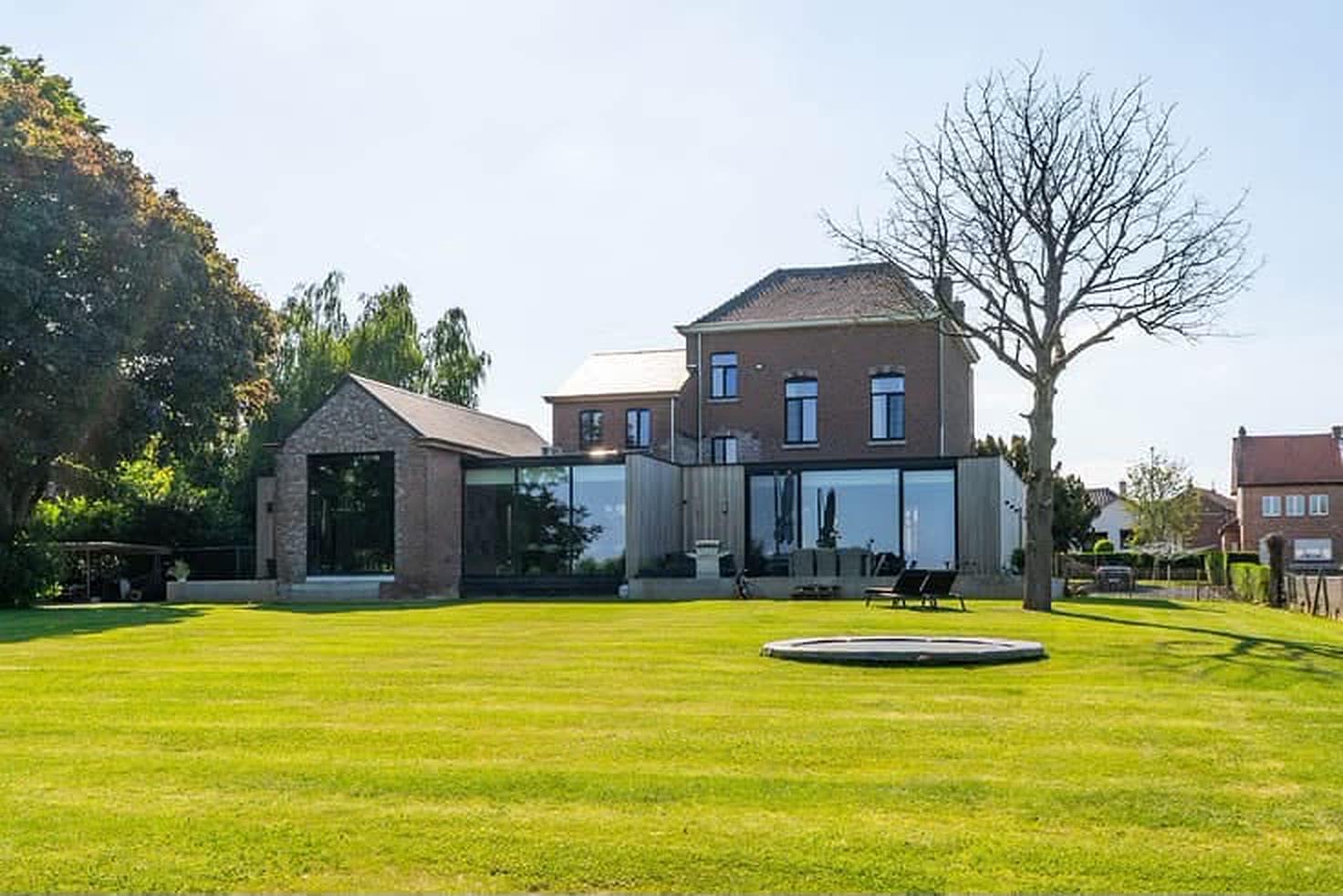Achterzijde van het domein met groot gazon, trampoline, kale boom en de glazen vleugel.